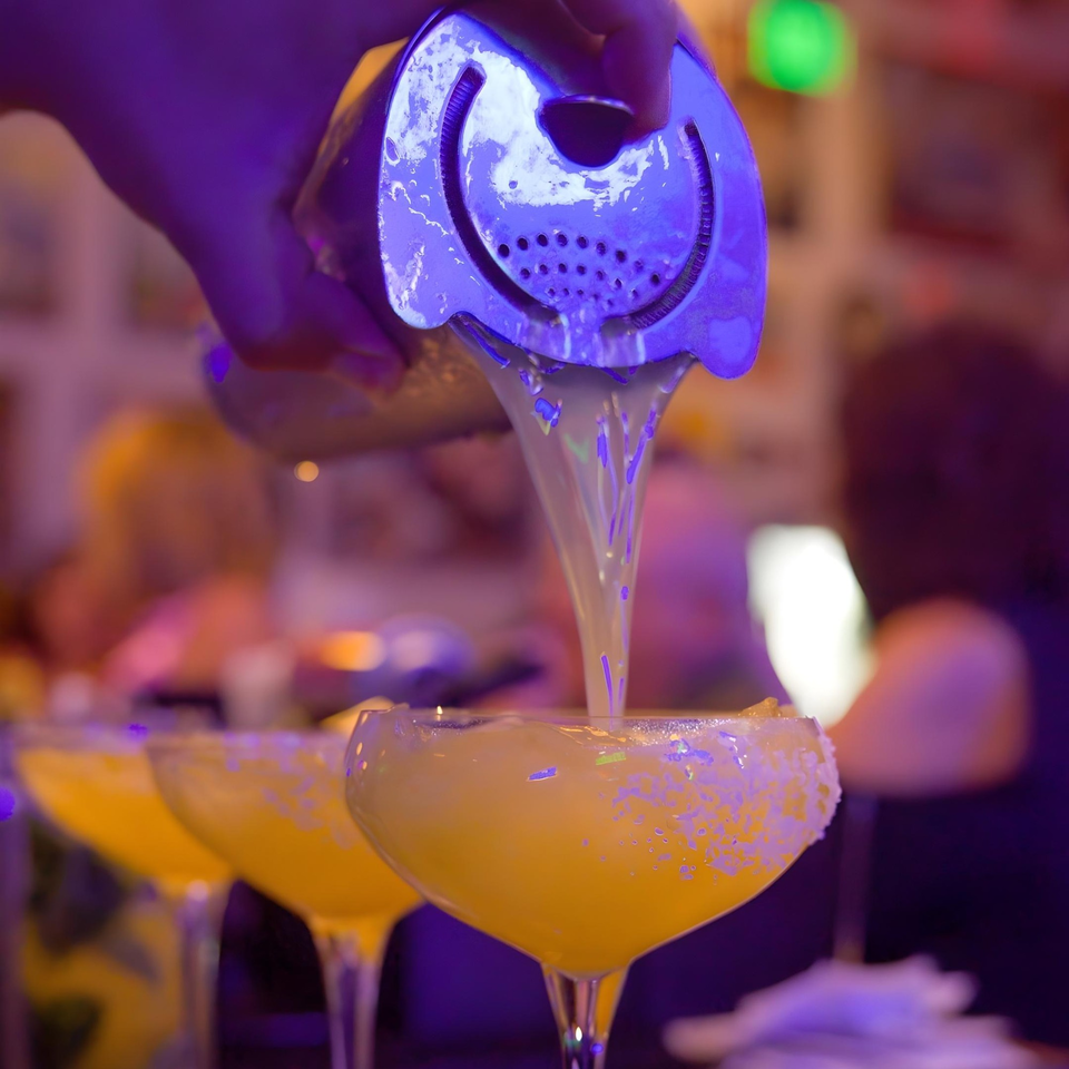 A bartender pours a vibrant yellow Cuban cocktail from a shaker into a sugar-rimmed glass.