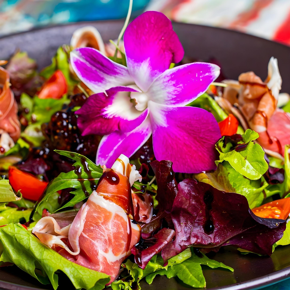 A vibrant Cuban salad with mixed greens, tomatoes, prosciutto, and a purple orchid garnish.