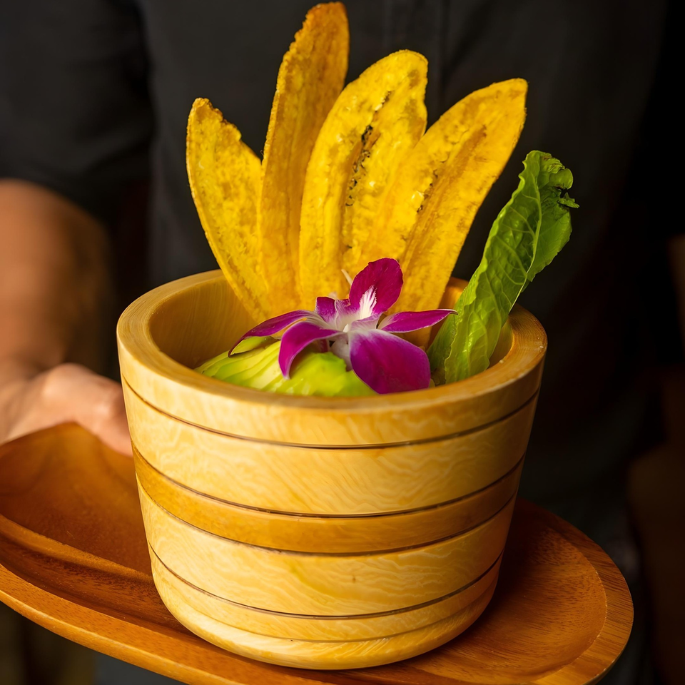 A wooden bowl of green avocado dip, topped with plantain chips and a purple orchid.