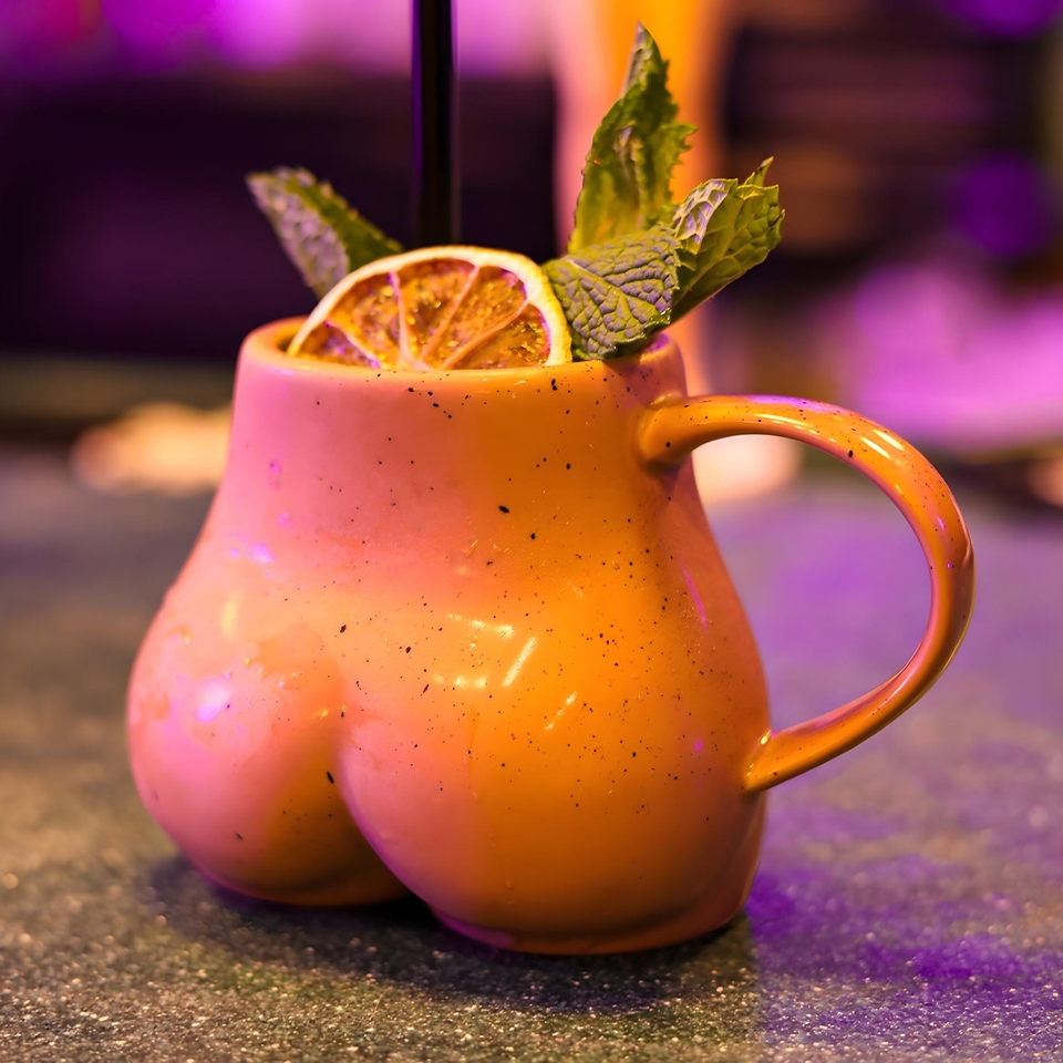 A vibrant orange cocktail in a unique, curvy mug, garnished with mint and a dried orange slice.